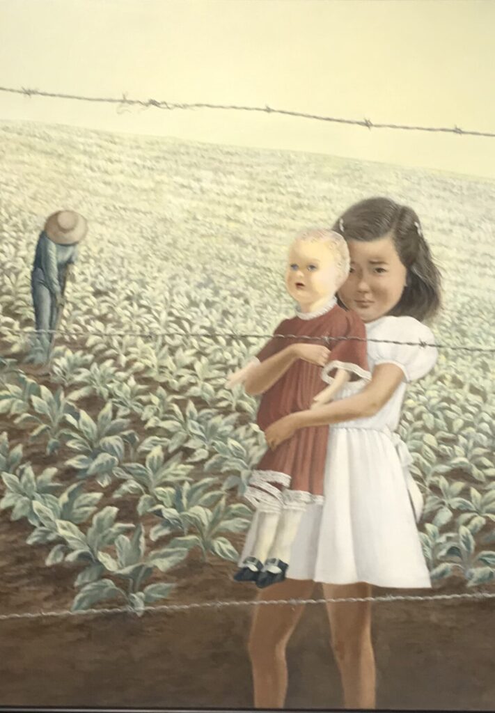 A small Japanese Canadian girl clutches her doll behind barbed wire looking as if she is about to cry while someone works the fields in the background.